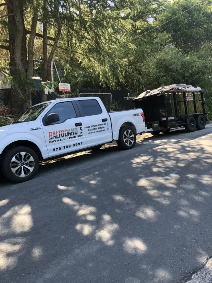 "A truck bearing the Baltodano's Drywall and Painting logo, parked on a road with a trailer attached, surrounded by trees."